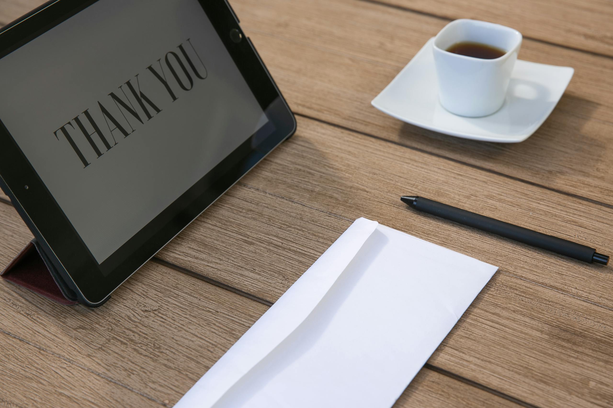 Sophisticated workspace setup with tablet displaying 'Thank You', cup of tea, pen, and envelope on wooden table.